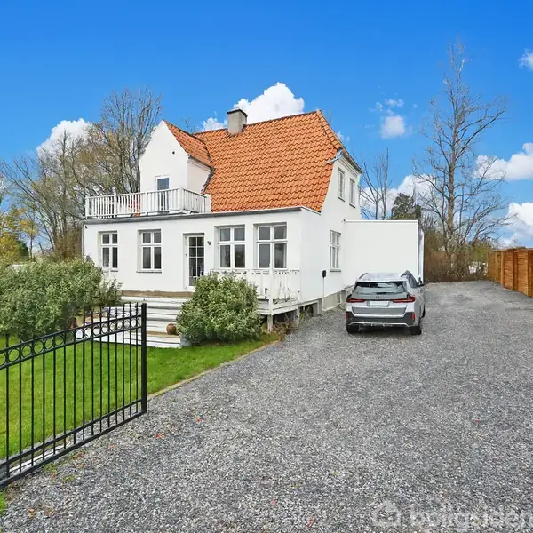 Et hvidt hus med rødt tag står ved en grusvej. En bil er parkeret ved siden af. Huset er omgivet af grønne buske og et sort hegn i et fredeligt område.