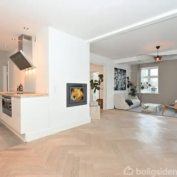 Stue og køkken i åben planløsning med trægulv og hvidmalede vægge. Køkkenet har en moderne emhætte og integrerede apparater. Stuen er møbleret med lænestole og dekoreret med planter.