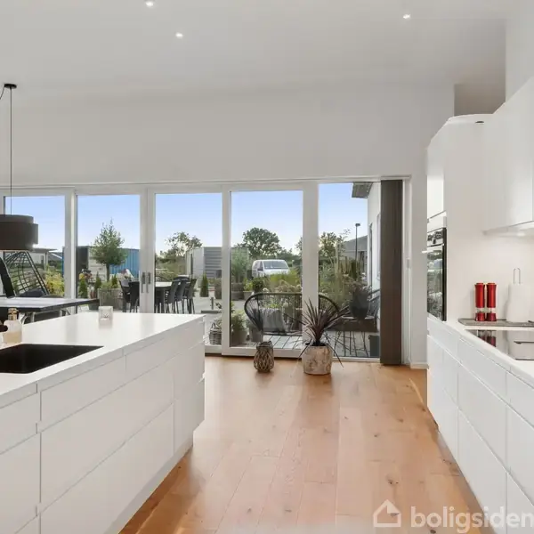 Et moderne, lyst køkken med hvide skabe og to sorte vaskarmaturer. Store vinduer i baggrunden giver udsigt til en terrasse og have. Ingen synlig tekst udover "boligsiden".