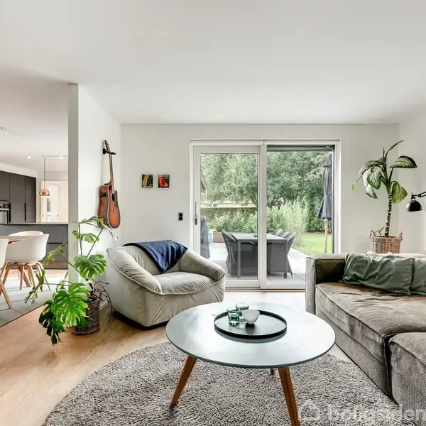 En stue med en grå sofa og lænestol står overfor et rundt sofabord. Store glasdøre fører til en have. Til venstre ses et spisebord og et køkken i baggrunden.