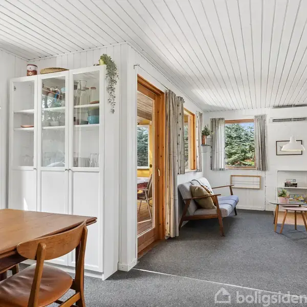 Et lyst stueområde med spisebord og stole i forgrunden, en tæppebelagt opholdszone med sofa og sofabord. Store vinduer og hvide vægge skaber en rummelig og lys atmosfære.