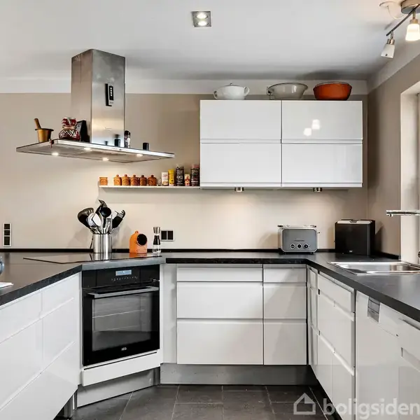 Hvidt, moderne køkken med sort bordplade, udstyret med ovn, emhætte, skåle og krydderier på hylderne. Store vinduer giver udsigt til have med efterårsfarver.