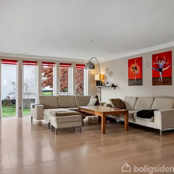 Sofagruppe i beige nuancer står rundt om et træbord i en lys stue med store vinduer, røde dekorationer og kunst på væggene; en lampeskærm hænger krummet over sofaen.