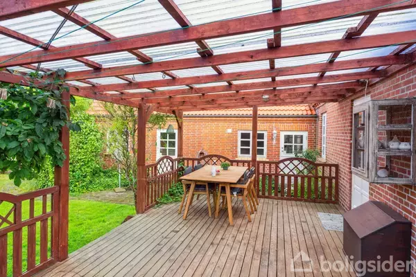 En træterrasse med et bord og stole står under et halvtag. Omgivet af en have og murstensbygning med vinduer.