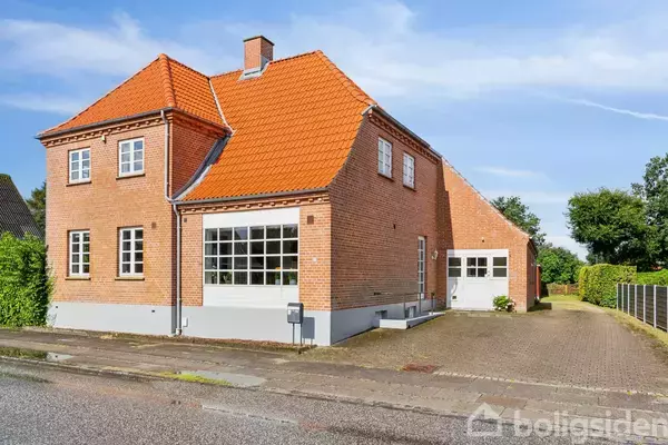 Murstenshus med rødt tegltag står stille langs en gade i et boligområde. Ved siden af huset ses en opkørsel, og omgivelserne består af hække og træer.