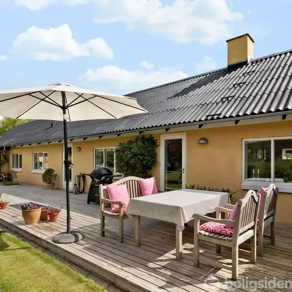 En terrasse med bord og stole under en stor parasol. I baggrunden ses et gult hus med sort tag omgivet af en velplejet have med grønne planter.
