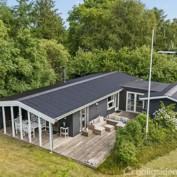 Sommerhus med sort tag og træterrasse ligger omgivet af tæt skov og græsplæne. Flisebelagt terrasse med udendørs møbler er synlig. Der er en flagstang foran huset.
