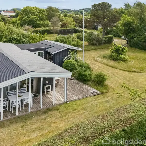 Et sommerhus med sort tag står på en trimmet græsplæne, omkranset af buske og træer. Under den overdækkede terrasse står et bord og stole.