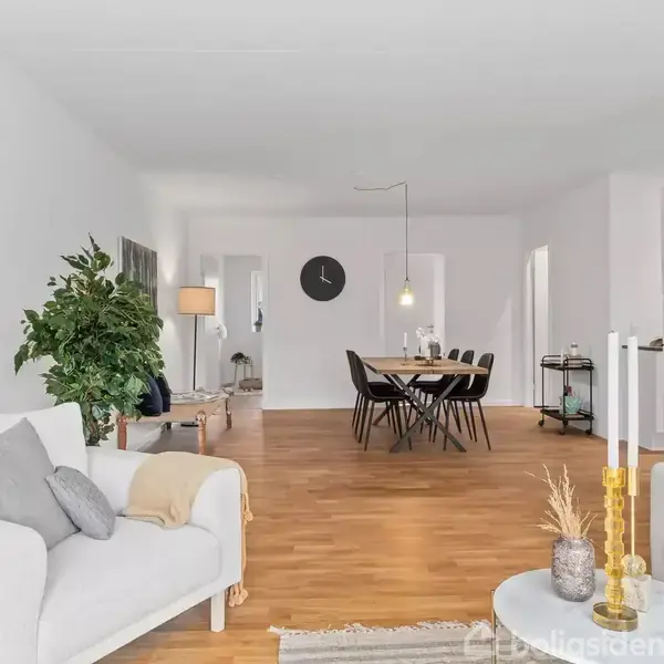 Et lyst stueområde med hvide vægge og trægulv. To sofaer rammer rummet, der indeholder et spisebord med seks stole. En vægklokke hænger centralt. Grønne planter og lamper skaber hygge.