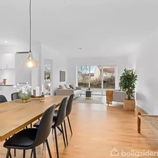 Spisebord af træ med sorte stole står i en stue. Flot lampe hænger over bordet. I baggrunden ses en sofa, store vinduer og planter. Væggene er hvide.
