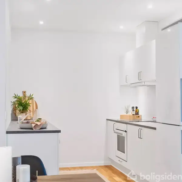Et moderne køkken med hvide skabe og en indbygget ovn. På bordet står krydderier, olier og dekorationer med planter og skærebrætter, der skaber en minimalistisk atmosfære.