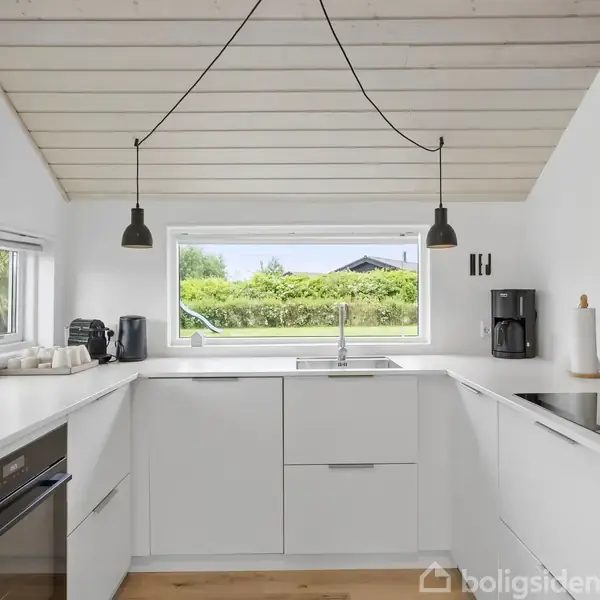 Køkkenvask i moderne, hvidt køkken med sort armatur; udstyret med hvidevarer; ovenlys af to sorte lamper; omgivet af store vinduer med udsigt til en grøn have.