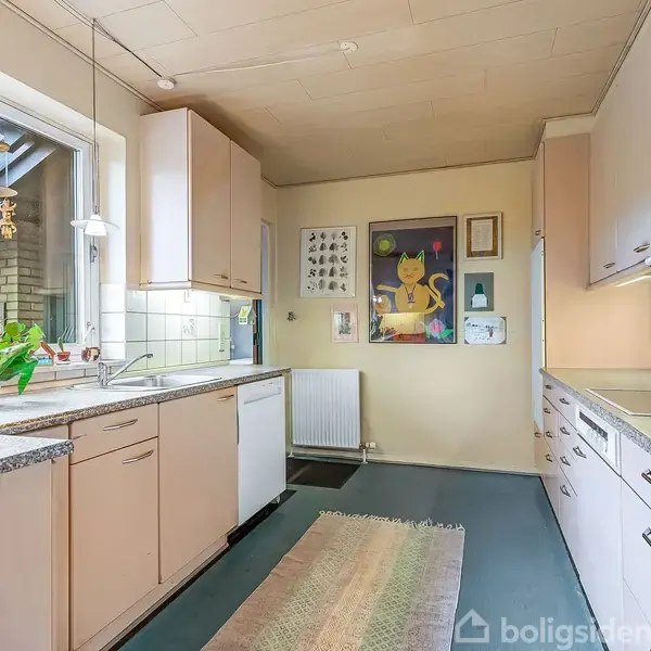 Et velbelyst køkken rummer lyse skabe, en ovn og vask med udsigt til et vindue. Dekorationer og planter pryder rummet, mens en tæppe løber langs det mørke gulv.