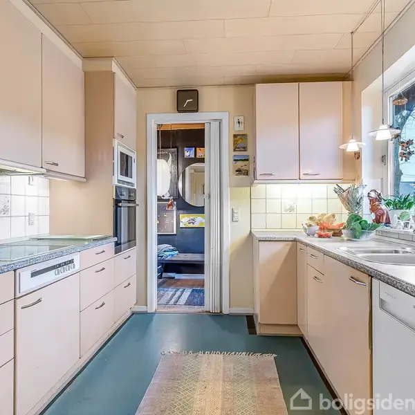 Køkken med lyse skabe og grå bordplader; opvaskemaskine og vask står under vinduet. Rum indrammet af hvide fliser og pastelfarvede skabe. En dør fører til et tilstødende rum.