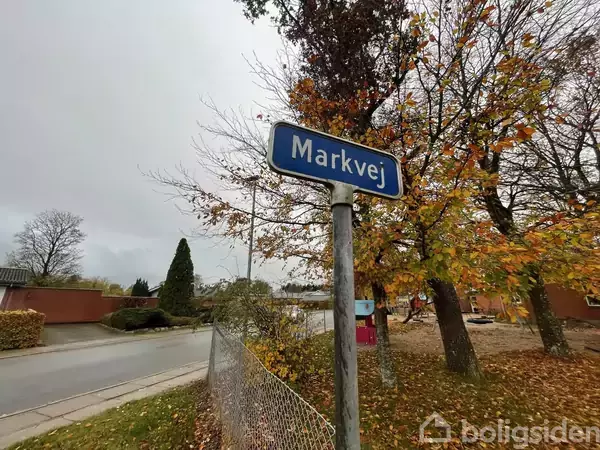 Et vejskilt viser teksten "Markvej". Det er placeret i en efterårslandskab med træer uden blade, et hegn og en vej til venstre.