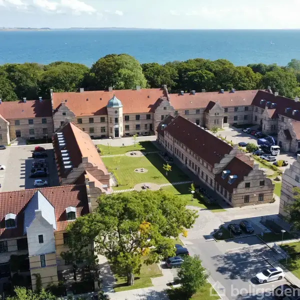 Et komplekst af gamle bygninger med røde tegltage omkranser en central gård. Nogle biler er parkeret, og området er omgivet af træer. Havet kan ses i baggrunden.