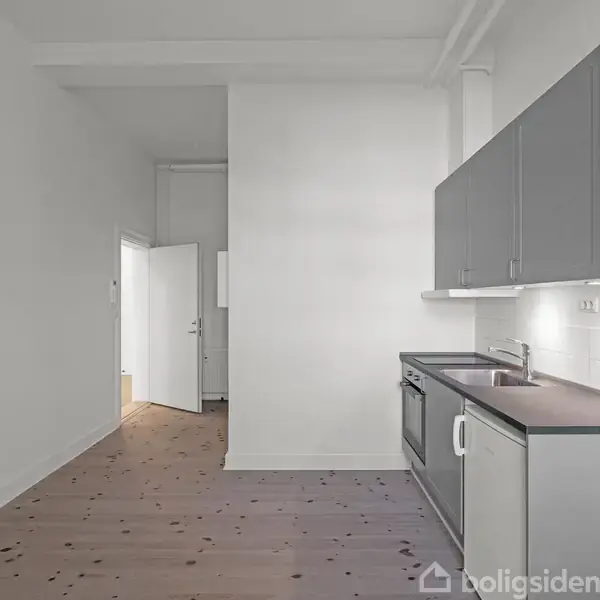 Et køkken med grå skabe og skuffer langs højre væg. Der er en vask og en ovn. På venstre side er et trælaminatgulv. I baggrunden ses åbne døre.