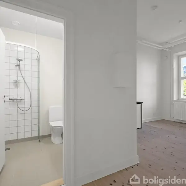 Et badeværelse med bruser og toilet ses gennem en åben dør. I tilstødende rum findes et vindue med udsigt til grønne træer.
