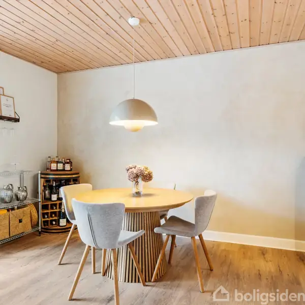 Rundt spisebord med tre stole står under en pendellampe i en spisestue med trægulv og træloft. En hylde med kasser og krukker ses ved siden af.