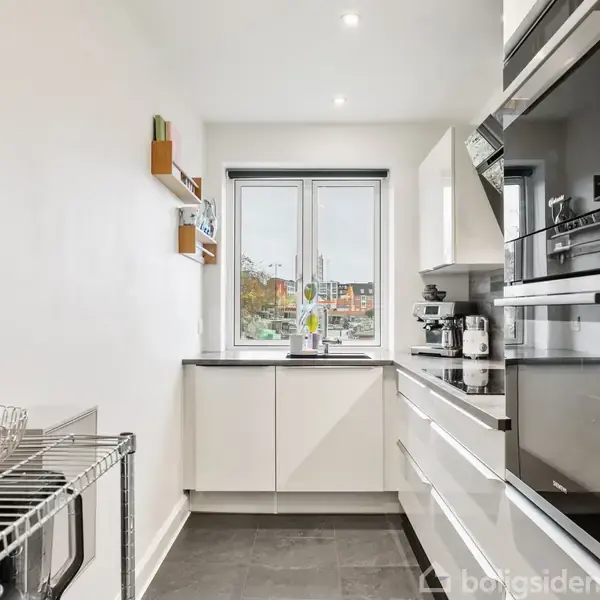 Et moderne køkken med hvide skabe og sorte apparater. En frugtskål står på en metalreol. Et vindue i baggrunden giver naturligt lys.