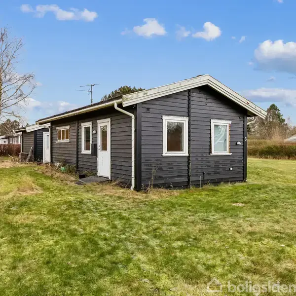 Sort træhus står stille på en grøn græsplæne under en skyfri blå himmel. Rundt omkring er der sparsom beplantning, herunder nogle nøgne træer og buske.
