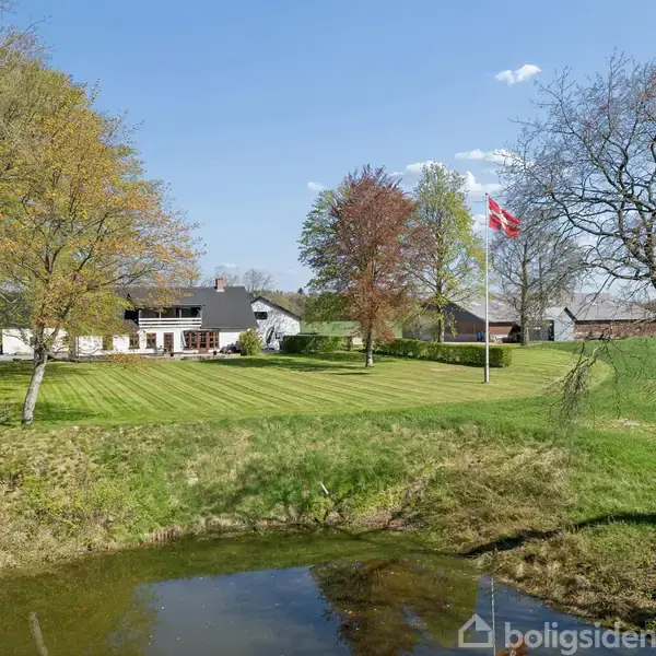Hus med sort tag og hvid facade står stille på en grøn plæne med træer og en dam, i baggrunden vejer Dannebrog på en flagstang i en grøn landskabssetting.