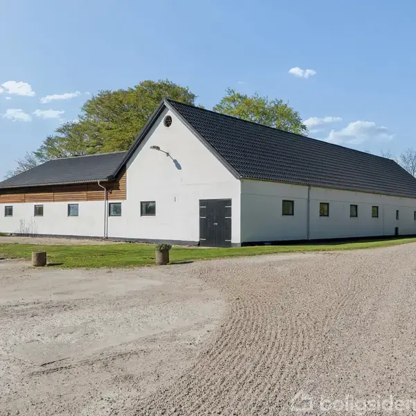 En stor, hvid landbygning med sort tag står på en grusvej, omgivet af grønne træer og en klar blå himmel i baggrunden.