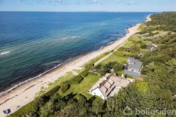 Hus ved kysten med stråtag ligger tæt ved stranden. Grønne buske og træer omgiver ejendommen, og den åbne blå havudsigt strækker sig mod horisonten i solrigt vejr.