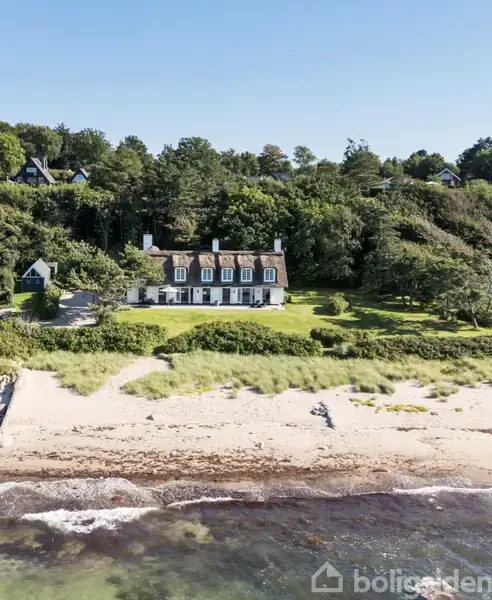 En stor, hvid bygning med flere kviste står stille på en frodig, grøn græsplæne omgivet af tæt skov. Foran er der en sandstrand med bølger, der rammer kysten.