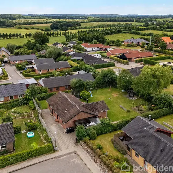 Luftfoto af et boligområde med huse omkranset af haver og grønne hegn, beliggende i et åbent landskab med marker og træer i baggrunden.