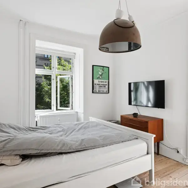 Soveværelse med en uopredt seng, en fladskærms-tv på væggen, og en trækommode. Store vinduer og glasdør åbner mod en balkon med haveudsigt.