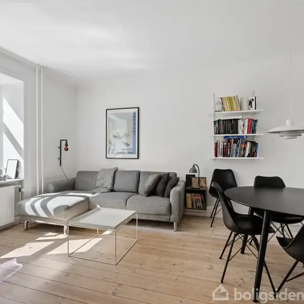 Grå sofa med puder står i et lyst stueområde. Ved siden af er der et rundt spisebord med sorte stole. Væggen har hylder med bøger og billeder.