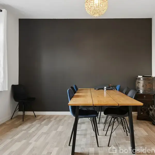 Et træbord med seks stole står i et rum med en sort væg og trægulv. Et stort vindue med hvidt gardin giver lys, og en rund lampe hænger i loftet.
