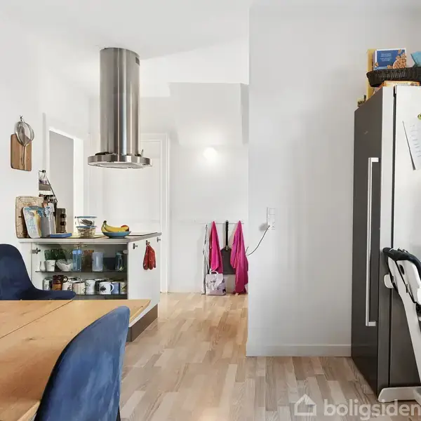 Spisebord med blå stole står i et lyst køkken. En emhætte hænger over en køkkenø med frugter. Til højre ses et køleskab og en barnestol. Desuden hænger jakker i baggrunden.