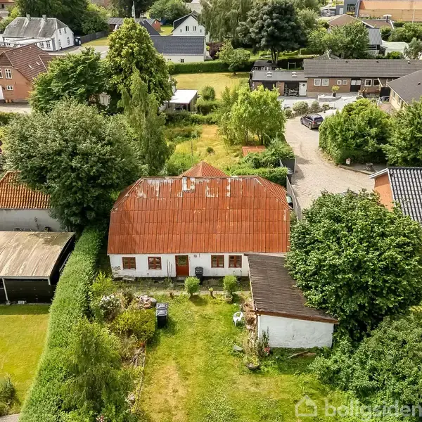 Rødt-tag-hus med hvid facade, omgivet af grøn have og store træer, beliggende i et boligområde med flere huse og veje i nærheden.