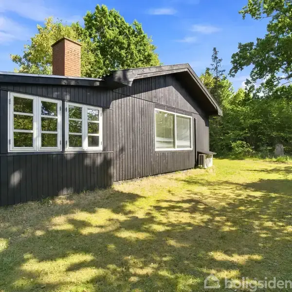 Et sortmalet træhus med hvidrammede vinduer står i en solrig have omkranset af grønne træer; en mur skorsten er synlig på taget.