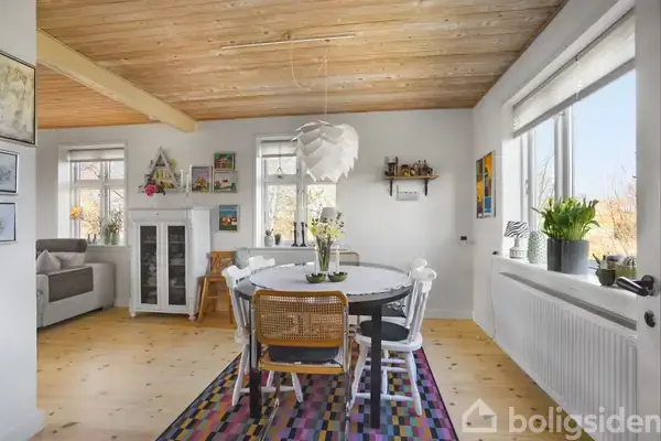 Rundt spisebord med hvide stole står på et farverigt tæppe. Ovenfra hænger en moderne lampe. Rummet har lyse vægge, trægulv og store vinduer, der bringer naturligt lys ind.