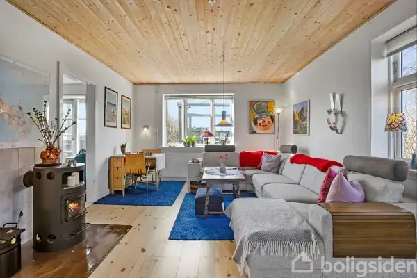 En stue med en grå sofa står ved siden af et sofabord, mens en brændeovn opvarmer rummet. Et spisebord med stole står i nærheden, omringet af lyse vægge og trægulv.