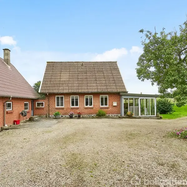Et rødt murstenshus med tegltag står stille ved siden af en stor gårdsplads. I højre side ses et grønt område med et stort, frodigt træ.