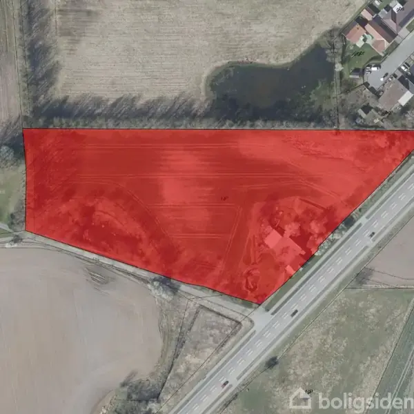 Rødt markeret område viser en trekantet grund ved en vej. Omgivet af marker og enkelte bygninger, ses området langs en hovedvej med felter og beplantning i nærheden.