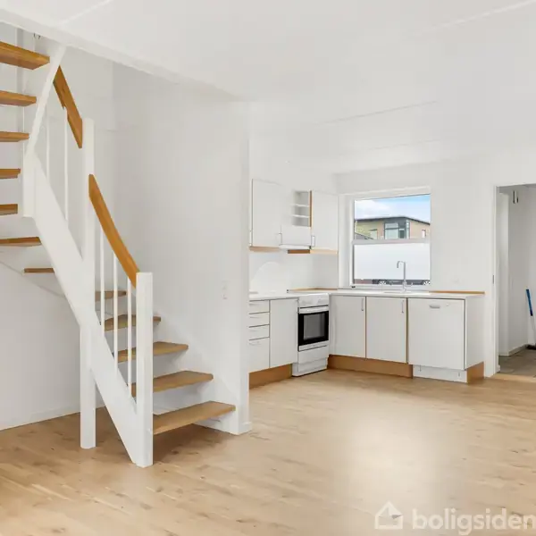 Trappe fører opad i et lyst rum. Køkken med hvide skabe i baggrunden. Døren fører til et tilstødende rum. Gulvet er af lyst træ.