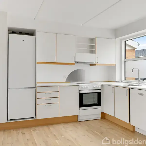 Et køkken med hvidt køleskab, komfur og opvaskemaskine. Køkkenet har hvide skabe og skuffer. En trætrappe ses til venstre. Et vindue giver udsigt til nabobygninger.