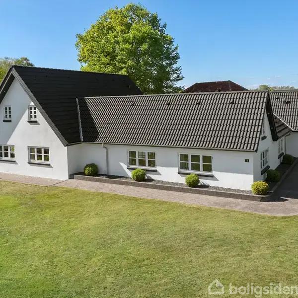 Hvid murstenshus med sort tag står i en velholdt have, omkranset af grønne buske og træer under en klar blå himmel. Græsplæne strækker sig foran huset.