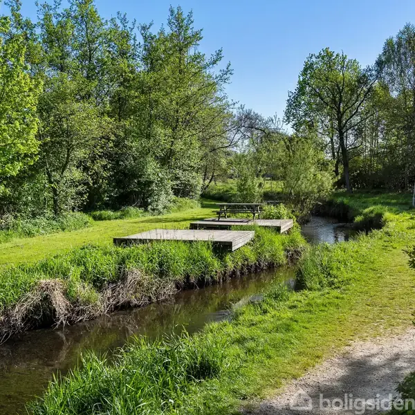 En smal bæk løber gennem et frodigt grønt område med træer og buske. To platforme, mulige picnicsteder, ses langs bredden i rolige omgivelser.
