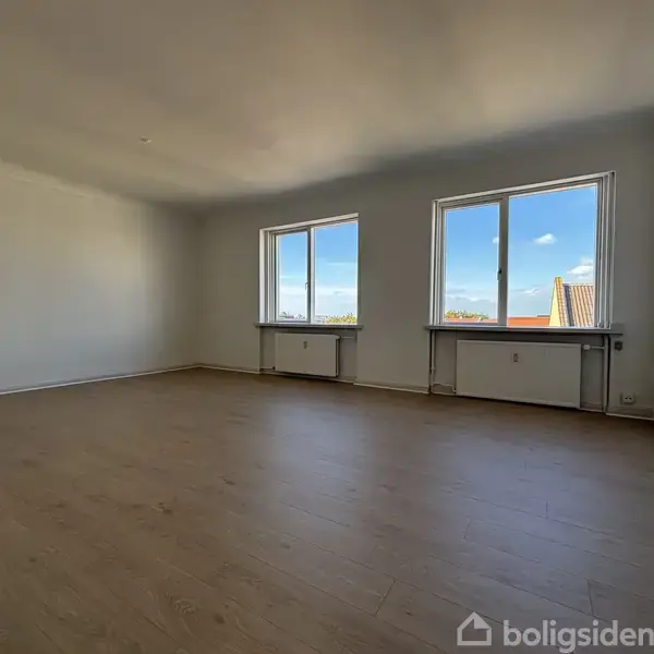 Et tomt værelse med lyse træplanker, to vinduer med udsigt til klare blå himmel og husstage, omgivet af hvide vægge og en lys, åben atmosfære.