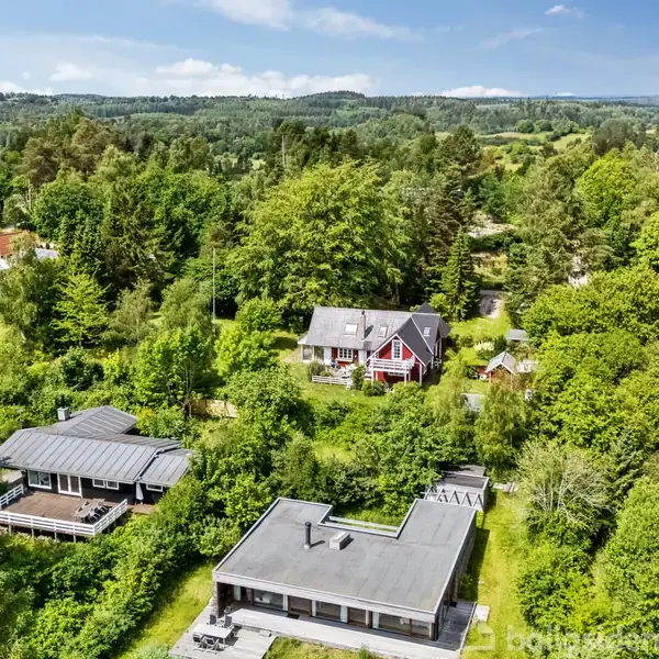 Huse er placeret blandt tæt bevoksede træer i en grøn, kuperet landskab. De omgivende haver bidrager til den landlige atmosfære under en blå himmel.
