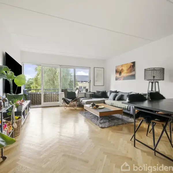 En stue med en stor sort sofa og sofabord i midten foran vinduer med udsigt til en altan. Der står en stor plante i venstre side, og et spisebord med stole er til højre. På væggen hænger kunstværker.
