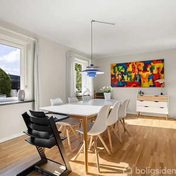 Spisebord omgivet af hvide stole står i et lyst rum med trægulv. En farverig abstrakt maleri hænger på væggen over en kommode. En blå lampe hænger over bordet.