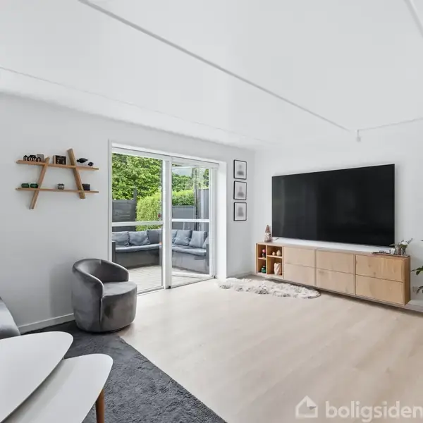 Stue med stor fladskærm på væggen og lys trægulv, sofa og stol til venstre, spisebord til højre. Store vinduer og dør fører ud til en terrasse med træmøbler.
