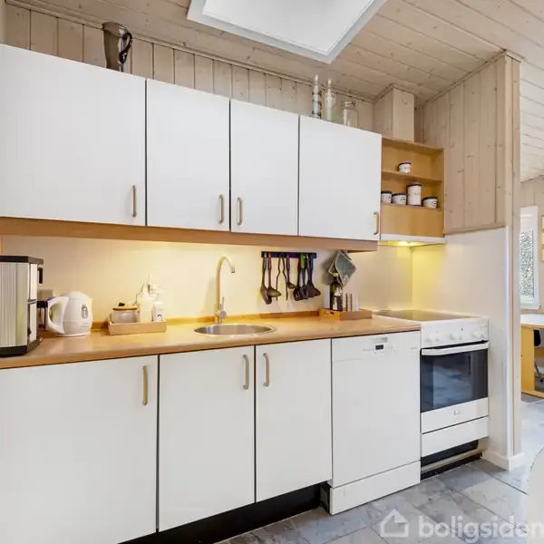 Et køkken med hvide skabe og en træbordplade; en elkedel og beholder står på bordet; gasblus ved siden af skrivebord med stol ved et vindue.